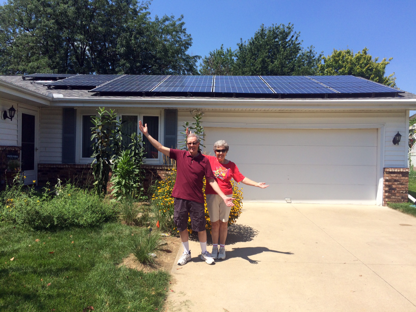 Two residents standing, arms outstretched, in front of their home with solar panels