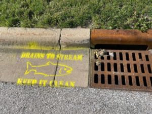 Example of a storm drain stencil. Reads, "Drains to stream. Keep it clean" in bright yellow spray paint to the left of a storm drain.